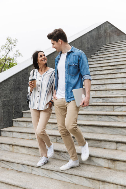Image of optimistic couple with paper cup and laptop talking while strolling down stairs outdoors - Foto, Bild