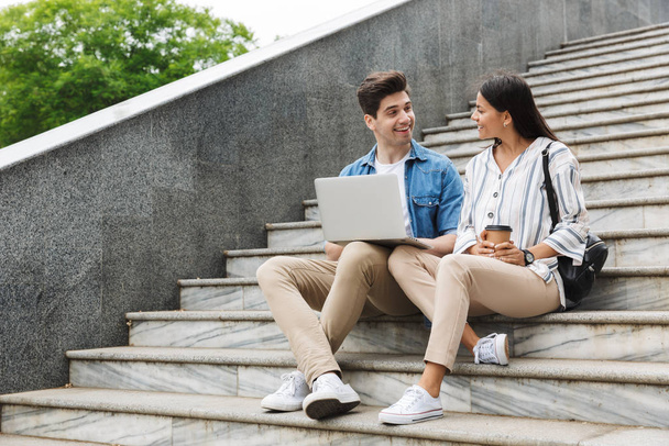 Image of kind couple with paper cup smiling and using laptop while sitting on stairs outdoors - Foto, imagen
