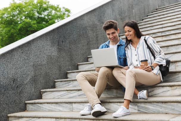 Image of optimistic couple with paper cup smiling and looking at laptop while sitting on stairs outdoors - Фото, изображение