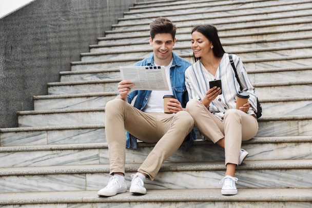 Happy young amazing loving couple business people colleagues outdoors outside on steps reading newspaper drinking coffee. - Foto, Imagen