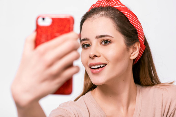 Closeup portrait of beautiful young brunette woman in casual clothes and red headband, taking self portrait over white studio wall. Happy female smiling and taking a selfie on her smart phone. - Foto, Imagen
