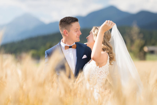 Bruidegom knuffels bruid teder terwijl wind waait haar sluier in tarwe veld ergens in Sloveense platteland. - Foto, afbeelding