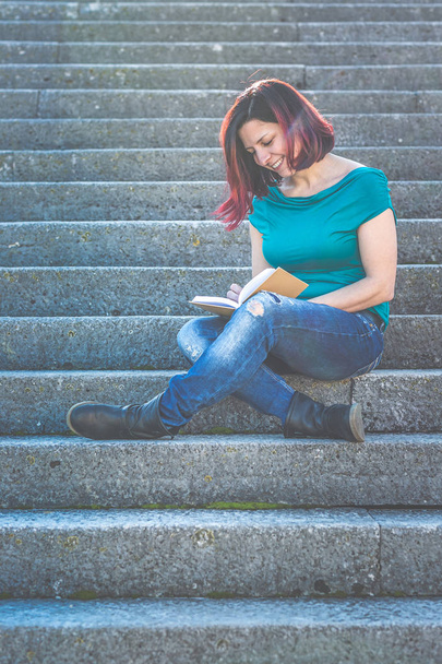 Merdivenlerde oturmuş kitap okuyan günlük kıyafetli bir kadın. - Fotoğraf, Görsel