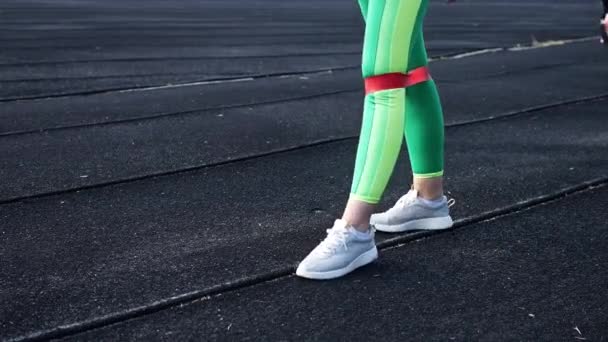 mooi jong meisje in groene sportlegging is bezig met een rubberen band voor fitness in het stadion. Maakt been oefening een elastische band voor fitness close-up - Video