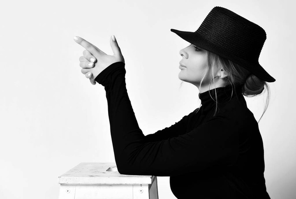 Black and white portrait in profile of blonde woman in black hat with brim doing a handgun gesture pointing at something - Photo, Image