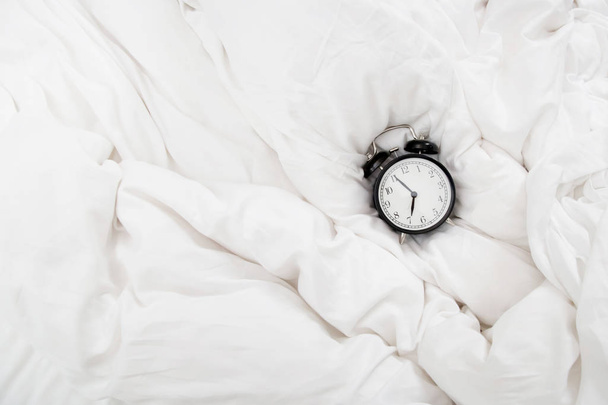 Relógio de alarme preto clássico nos lençóis brancos enrugados
 - Foto, Imagem