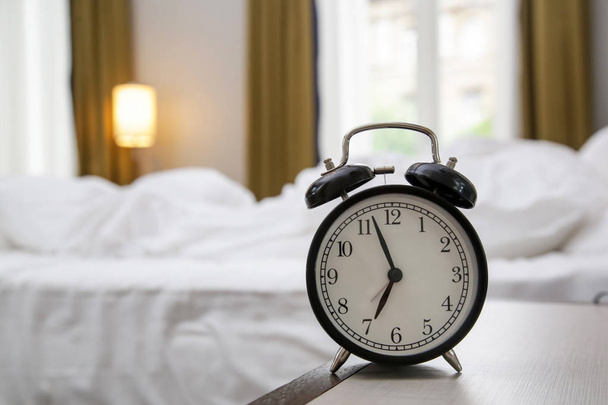 Relógio de alarme preto clássico na mesa de cabeceira mostrando sete horas com um quarto no fundo
 - Foto, Imagem