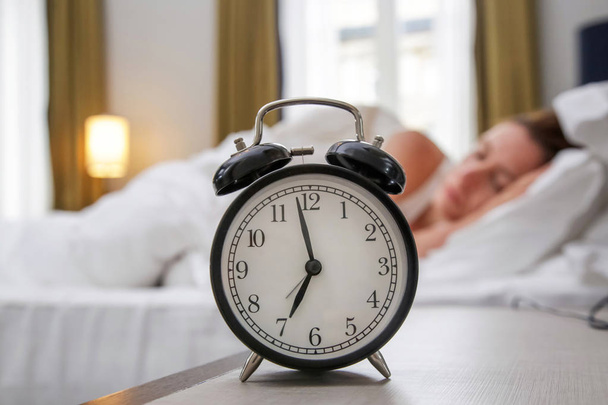 Klassieke zwarte wekker op de nachtkastje die zeven uur toont met een vrouw die op de achtergrond slaapt - Foto, afbeelding