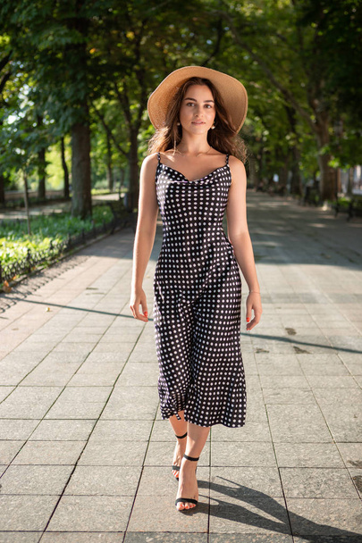 Beautiful young brunette woman dressed in a black dress and a hat with wide flaps takes a walk in a park during warm summer day enjoying sunlight. - Фото, изображение