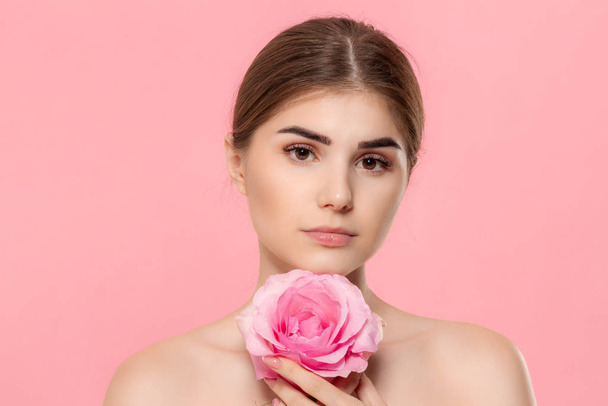 ピンクの背景の上に孤立した顔に近いピンクのバラの花を持つカメラを見て美しい若い女の子のクローズアップポートレート. - 写真・画像