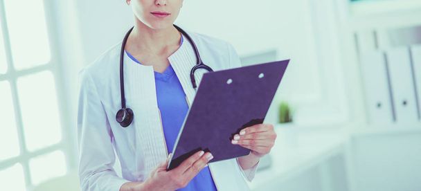 Молодая улыбающаяся женщина-врач со стетоскопом держит папку в кабинете врача - Фото, изображение