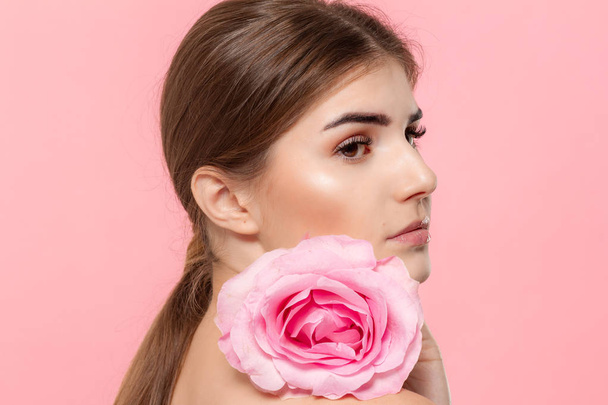 Κοντινό στο πορτρέτο ενός όμορφου νεαρού κοριτσιού με ροζ τριαντάφυλλο που απομονώνεται πάνω από ροζ φόντο. - Φωτογραφία, εικόνα