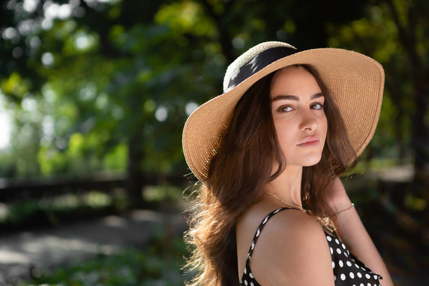 Ritratto ravvicinato di una bella giovane donna bruna vestita con un vestito nero e un cappello con ampie falde fa una passeggiata in un parco durante la calda giornata estiva godendo della luce del sole
. - Foto, immagini