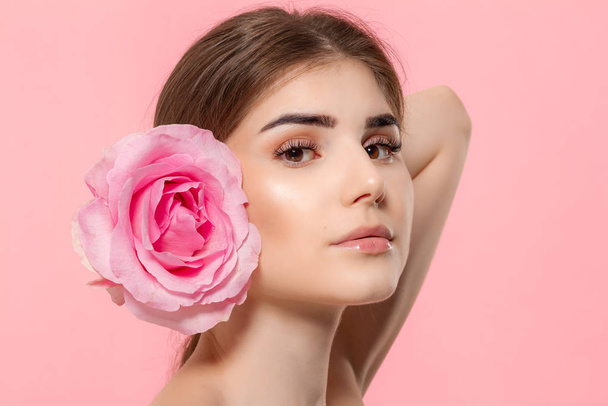 Close-up portrait of a beautiful young girl looking at the camera holding pink rose flower close to face isolated over pink background. - Photo, Image