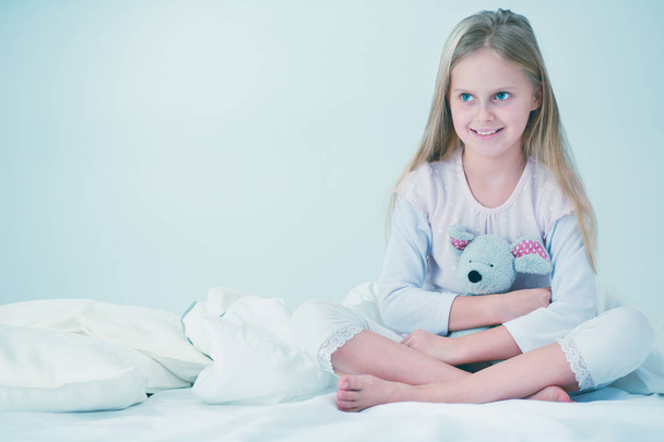 Küçük kız yatak odasında yatağın üzerine oturuyor. Küçük kız yatakta oturuyor ve bir pijama giymiş . - Fotoğraf, Görsel