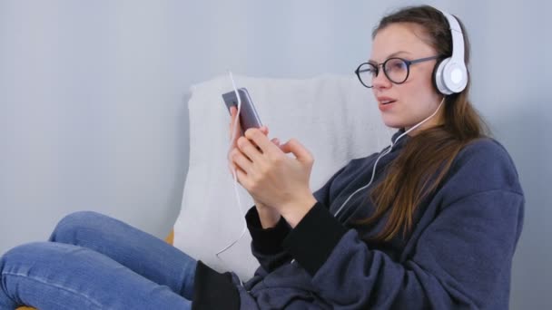 Woman in glasses listens music in headphones on smartphone and sings a song sitting in armchair. - Záběry, video