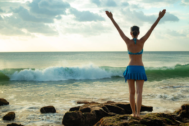 Emocionada joven levantando brazos en la playa frente al océano. Vista desde atrás. Puesta de sol en la playa. Bali, Indonesia
. - Foto, Imagen