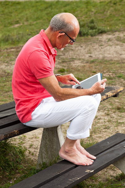 Пенсионер отдыхает и пользуется планшетом за столом в парке
 - Фото, изображение