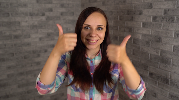 A beautiful emotional woman in a plaid shirt laughs happily and shows thumbs up. A woman shows a gesture of approval on a brick background. The concept of like or dislike. - Footage, Video