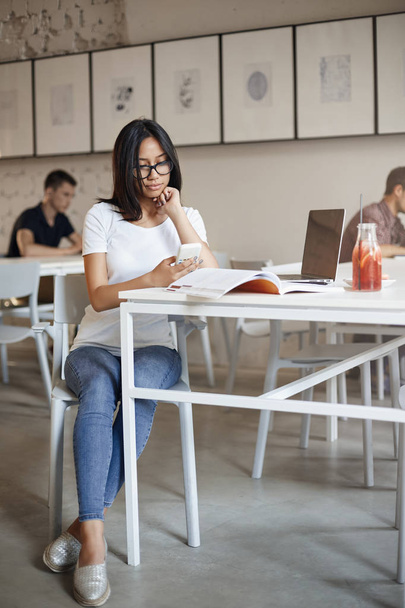 Függőleges shot elfoglalt fiatal nő a szemüveg ül egyedül kávézó körül a munkaanyag notebook és laptop, smartphone segítségével, görgetés szociális média feed alatt szünetet tanul - Fotó, kép