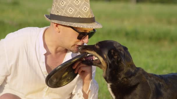 Zeitlupe, Nahaufnahme: Ein fröhlicher kaukasischer Mann küsst und spricht mit seinem fröhlichen Hund, der sein Spielzeug-Frisbee in der Hand hält, auf einen lustigen Ausflug ins Grüne. Herrchen teilt schöne Momente mit seinem entzückenden Haustier. - Filmmaterial, Video