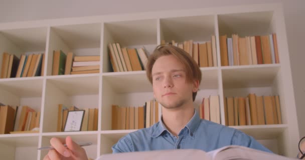 Closeup portrait of young attractive caucasian male student in glasses learning being thoughtful in library indoors with bookshelves on background - 映像、動画