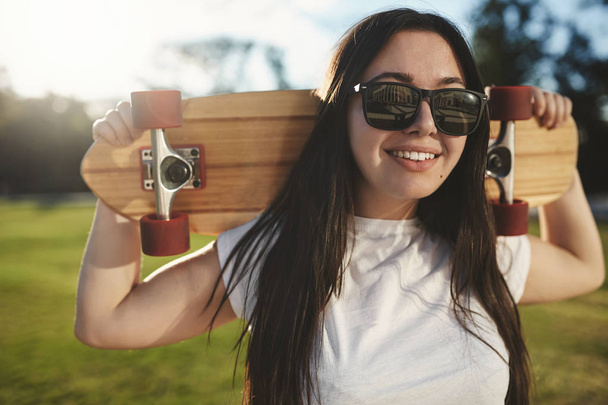 Close-up carefree relaxed skater girl chilling on grass at sunny park, enjoy summertime, wear sunglasses, white t-shirt, hold wooden penny board behind head, smiling joyfully - Foto, imagen
