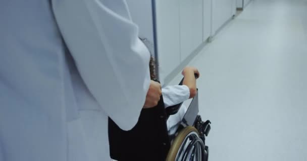 Close-up of Caucasian male doctor pushing pushing African american boy in wheelchair in hospital. They are passing through corridor 4k - Кадры, видео