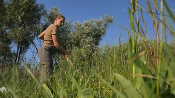 Jonge vrouw maaien het groene gras met de zede op het zomer veld aan de rivier. Prachtig landschap. Werk in het veld. Echte landelijke vrouw. - Video