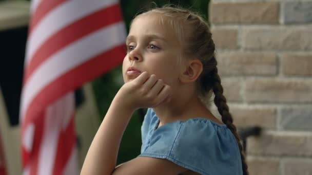 Sad girl missing father standing house balcony, american flag background patriot - Кадры, видео