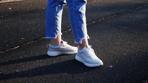 Close-up of the legs of a young white girl in white sports trainers and blue jeans who poses on camera shows her trainers standing on a black sports court. Shoe posing in the frame - Materiał filmowy, wideo