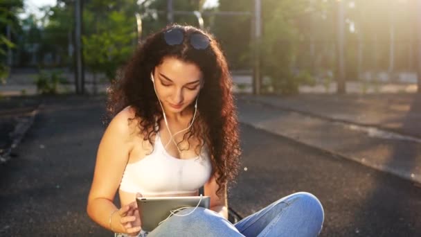 Young, happy, smiling caucasian girl with long curly hair, black sunglasses, white top and blue jeans sits on sports court wearing headphones, laughs, listens to music and texts on phone amid summer sunset - Footage, Video