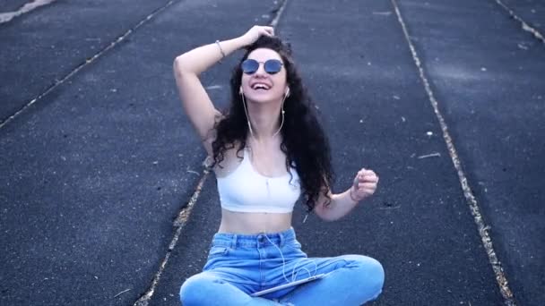 A young, happy, smiling caucasian girl in black sunglasses, a white top and blue jeans sits on a sports court wearing headphones listening to music and adjusting her long black curly hair - Materiał filmowy, wideo