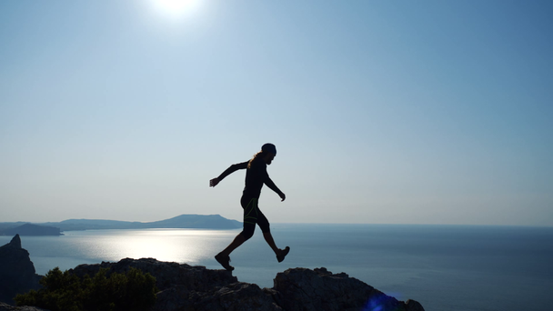 Young long-haired inspired man walks by a sharp mountain ridge towards the summit above the sea against beautiful blue sky at sunrise. Silhouette of a confident happy hiker man standing on the summit. - Footage, Video