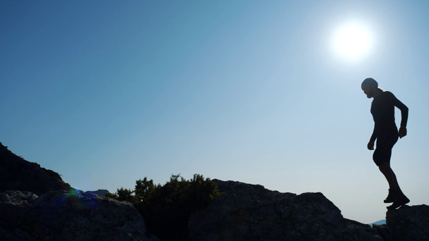 Jonge langharige geïnspireerde man verheft zijn handen op de top van een berg boven de zee tegen de prachtige blauwe hemel. Silhouet van een gelukkige wandelaar die op de top staat. - Video