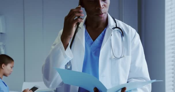 Front view of African American male doctor talking on mobile phone in hospital. He is looking at medical file 4k - Imágenes, Vídeo