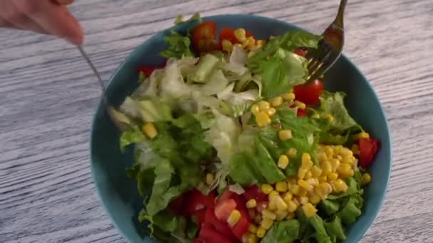 A woman stirs two forks a vegetable salad with tomatoes, herbs and corn in an aquamarine plate. On a wooden painted white table. - Video