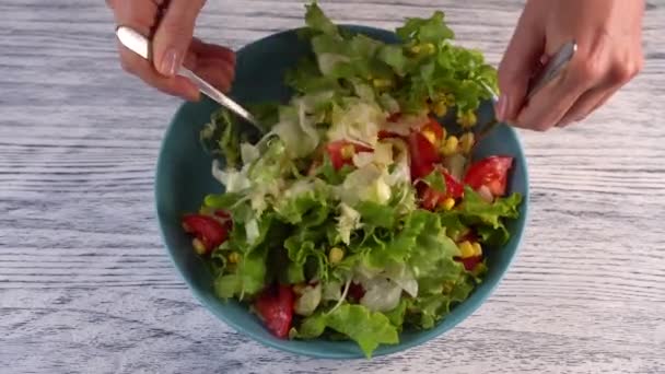 Una mujer revuelve dos tenedores una ensalada de verduras con tomates, hierbas y maíz en un plato de aguamarina. Sobre una mesa blanca pintada de madera
. - Imágenes, Vídeo