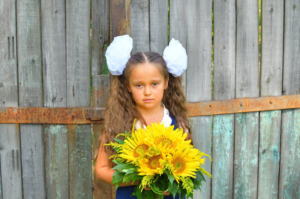 Portrét krásného mladého prvního gradera s kyticí slunečnic v slavnostní školní uniformě na pozadí starého dřevěného plotu. - Fotografie, Obrázek