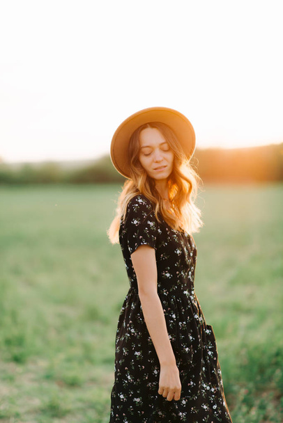 Young, Gentle Girl in hat Walks Through A green Meadow In The Rays Of The Sunset - Fotografie, Obrázek