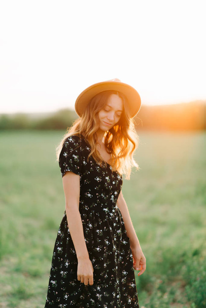 Joven, Chica gentil en sombrero Camina a través de una pradera verde en los rayos de la puesta del sol
 - Foto, Imagen