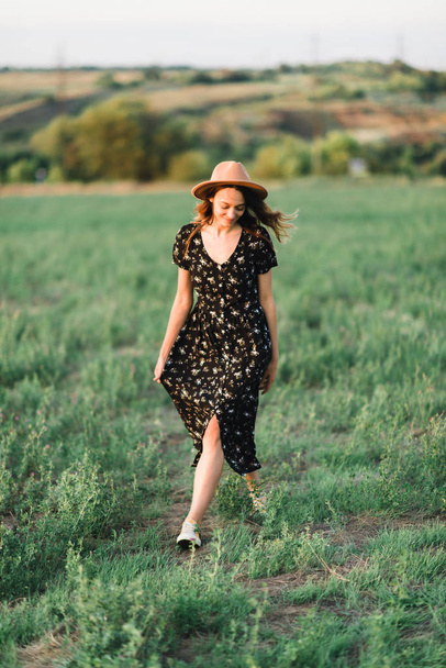 Fiatal, szelíd lány kalapban sétál át A zöld rét a sugarai a naplemente - Fotó, kép