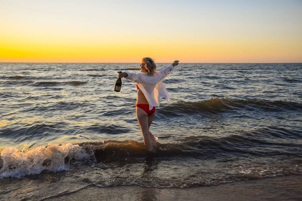 Chica joven en la playa del mar con una botella de champán y una copa, la mujer entró en el agua en la playa
. - Foto, Imagen