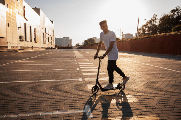Young guy dressed in jeans and t-shirt is riding a scooter on the square paved with tiles near the building on the sunny day - Фото, зображення