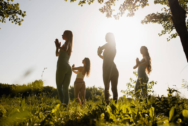 Four young women are practising yoga in the open air standing with folded hands near the chest in the nature on the grass on the sunny day - Foto, imagen