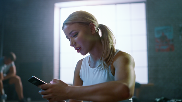 Zelfverzekerd mooie atletische jonge vrouw is met behulp van een smartphone terwijl zittend op een verdieping in een loft Gym. Ze typt een boodschap en denkt. Een man oefeningen op de achtergrond. - Video