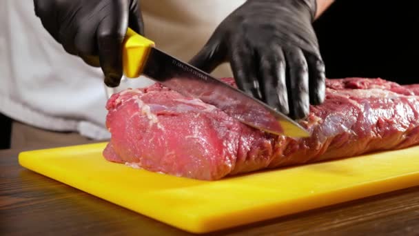 Siyah eldivenli profesyonel bir kasap keskin bir bıçakla entrecote biftek için çiğ et bir parça dilimler. Aşçı ızgara üzerinde yemek için bir hazırlık yapar. - Video, Çekim