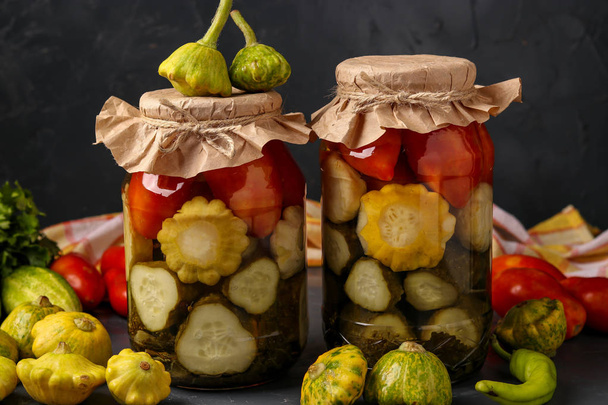 Surtido de pepinos en escabeche, patissons y tomates en frascos sobre un fondo oscuro, orientación horizontal
 - Foto, Imagen