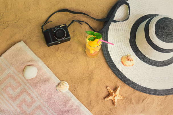 Καλοκαιρινές διακοπές στην παραλία. Φωτογραφία φωτογραφική μηχανή, καπέλο, καλοκαιρινό ποτό, πετσέτα, αστερίες και κοχύλια στην παραλία άμμου. Επιλεκτική εστίαση. - Φωτογραφία, εικόνα