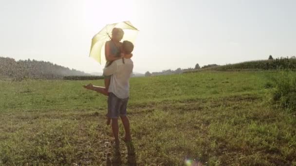 SLOW MOTION, LENS FLARE: Lovely young boyfriend and girfriend dance in the cool summer rain during their romantic date in the sunny green meadow. Man and woman in love spin in the spring rainfall. - Felvétel, videó